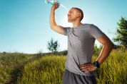 Ein Mann in grauer Sportkleidung erfrischt sich nach dem Training, indem er sich Wasser aus einer Plastikflasche über Gesicht und Kopf giesst. Er trägt ein graues T-Shirt und Shorts sowie eine dunkle Sportuhr am Handgelenk. Die Szene spielt sich in einer natürlichen Landschaft mit hohem Gras und einzelnen Bäumen ab, unter einem klaren blauen Himmel und. Die Sonne befindet sich hinter dem Kopf des Mannes und suggeriert Hitze.   Ein Mann in grauer Sportkleidung erfrischt sich nach dem Training, indem er sich Wasser aus einer Plastikflasche über Gesicht und Kopf giesst. Er trägt ein graues T-Shirt und Shorts sowie eine dunkle Sportuhr am Handgelenk. Die Szene spielt sich in einer natürlichen Landschaft mit hohem Gras und einzelnen Bäumen ab, unter einem klaren blauen Himmel und. Die Sonne befindet sich hinter dem Kopf des Mannes und suggeriert Hitze.